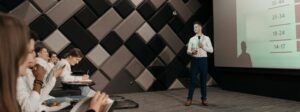 Homem de terno presidindo uma palestra em frente à uma projeâo com dados